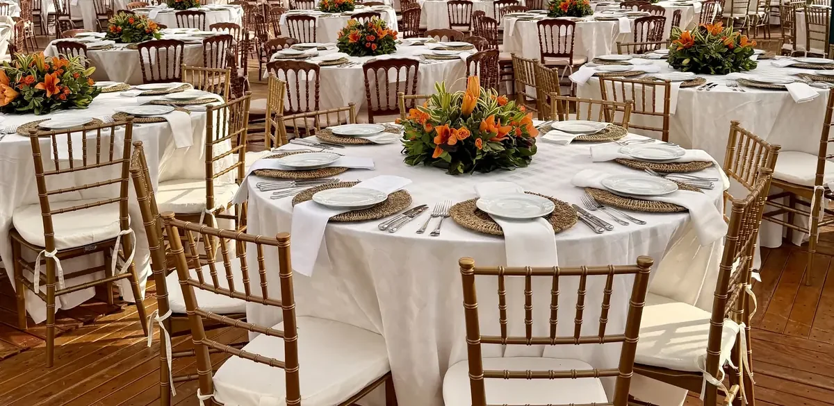 Mesas redondas de evento corporativo montadas sobre piso de madeira, com toalha branca, sousplats de fibra, taças, talheres e cadeiras douradas de encosto ripado