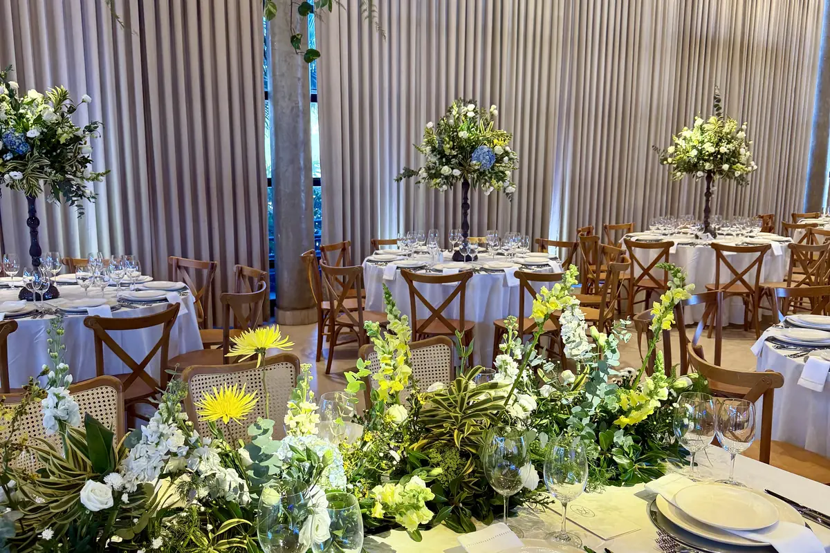 Mesa longa montada em salão, com caminho de flores brancas e amarelas no centro, e ao fundo mesas redondas de toalha branca com cadeiras de madeira e arranjos altos
