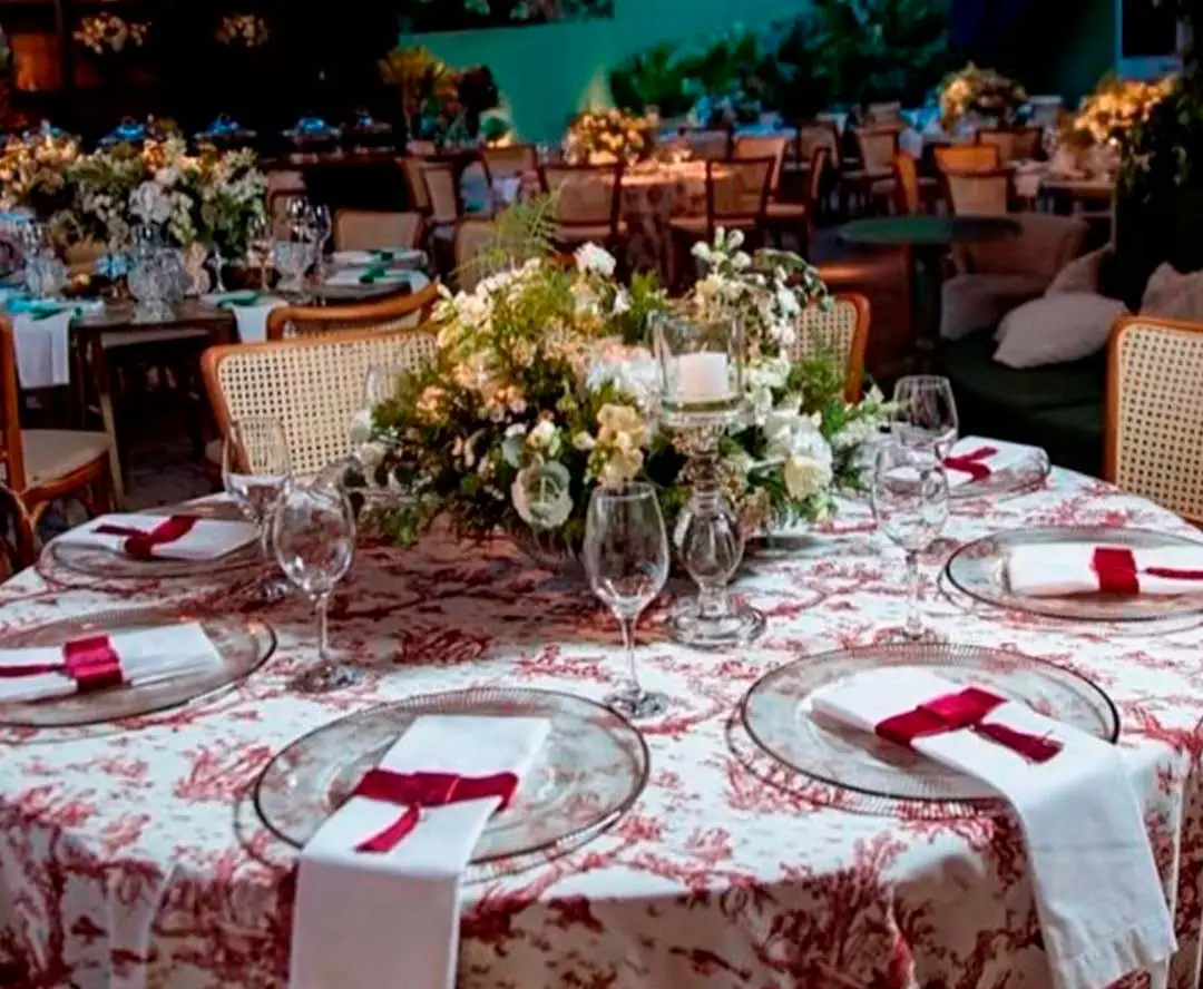 Mesa redonda com toalha Toile de Jouy bordô, sousplat de cristal, guardanapos brancos com laço bordô e arranjo de flores no centro