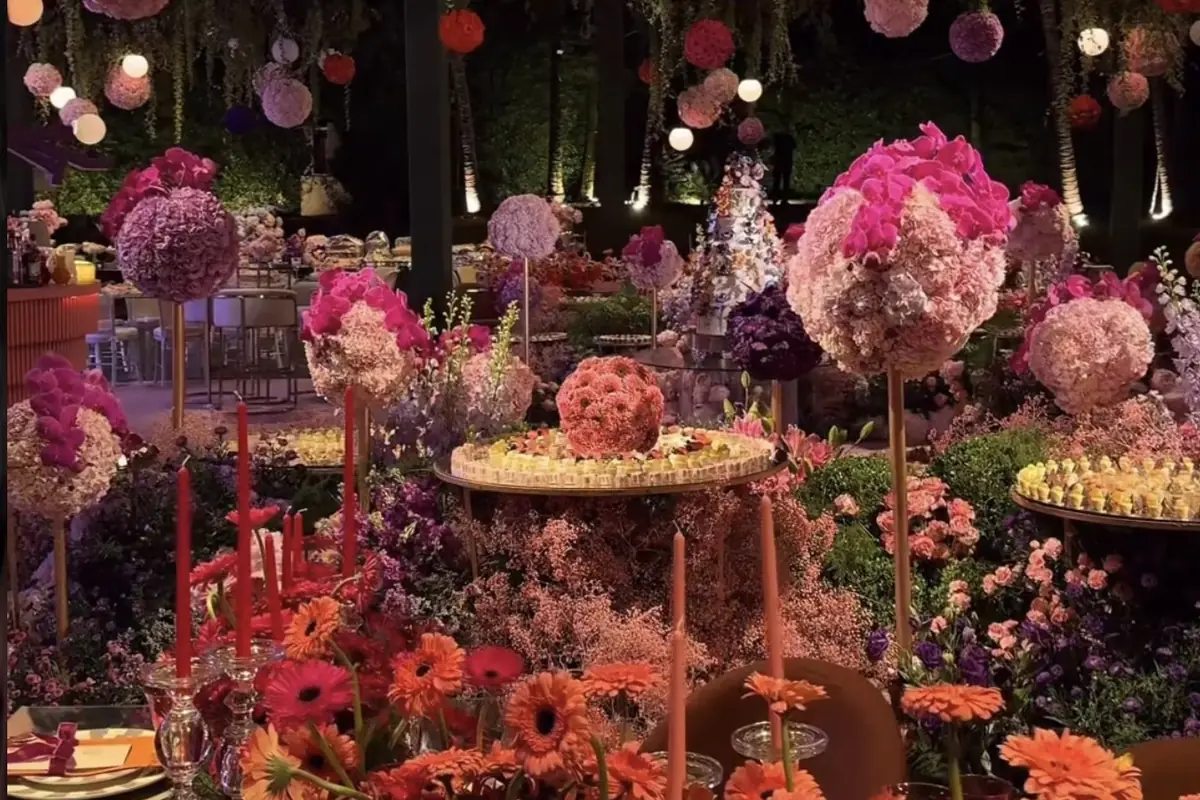 Mesa de doces com arranjos altos de flores em tons de rosa e laranja, boleiras e suportes redondos, sob luminárias de papel penduradas