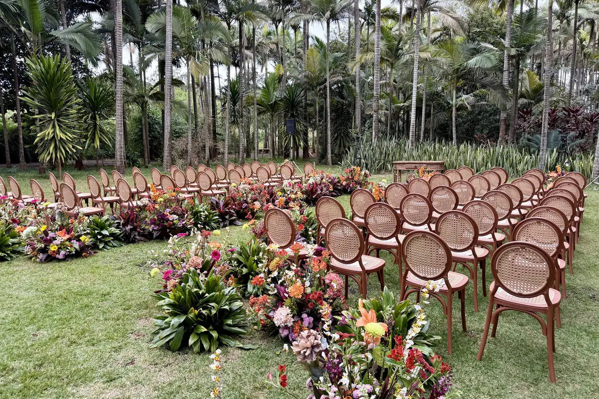 Cerimônia montada no gramado, com cadeiras de madeira em duas alas e canteiros de flores tropicais coloridas no corredor