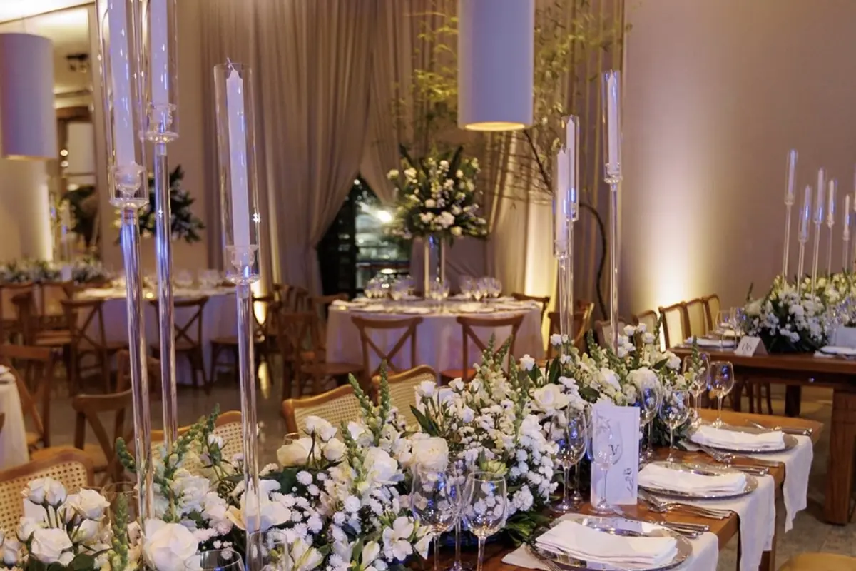 Mesa longa de jantar posta em salão, com caminho de flores brancas no centro, castiçais altos de cristal e guardanapos com menu sobre os pratos