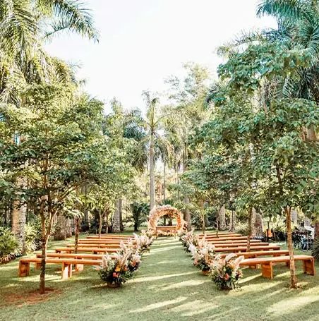 Cerimônia em jardim com bancos de madeira de demolição dispostos em duas alas, arranjos de flores nas pontas e arco de flores ao fundo