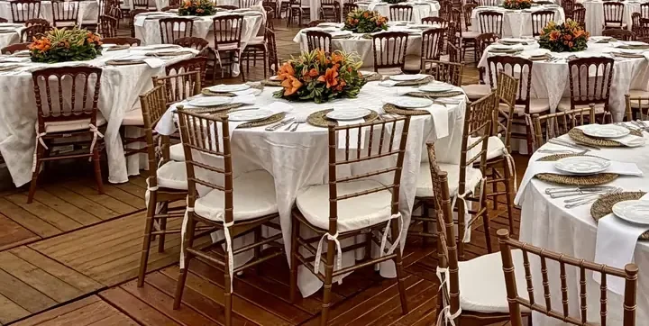 Vista ampla de pavilhão montado para evento, com mesas redondas de toalha branca em série, cadeiras douradas e de madeira escura e arranjos de flores laranja ao centro de cada mesa, sobre piso de madeira