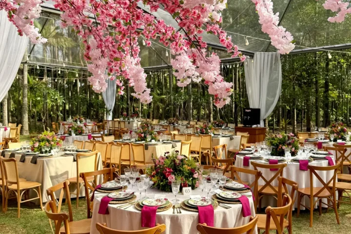 Recepção sob tenda transparente no jardim, com galhos de flores rosa suspensos, mesas redondas de toalha creme, guardanapos pink e cadeiras de madeira