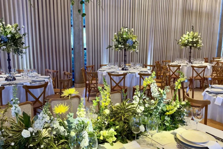 Mesa longa montada em salão, com caminho de flores brancas e amarelas no centro, e ao fundo mesas redondas de toalha branca com cadeiras de madeira e arranjos altos